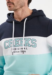 Hoodie z kapturem z wzorem w kolorze granatowym, białym i tealowym. Zawiera grafikę "CREEKS" oraz sznurki z metalowymi końcówkami. Wykonany z miękkiego materiału.