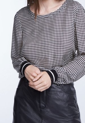 Femme portant un haut à manches longues noir et blanc à motif pied-de-poule avec des poignets noirs et une jupe en cuir noire, les mains jointes à la taille.