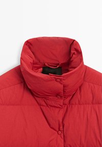 Röd vadderad jacka i närbild med hög krage, med slät textur, rundade knappar och enkel design.