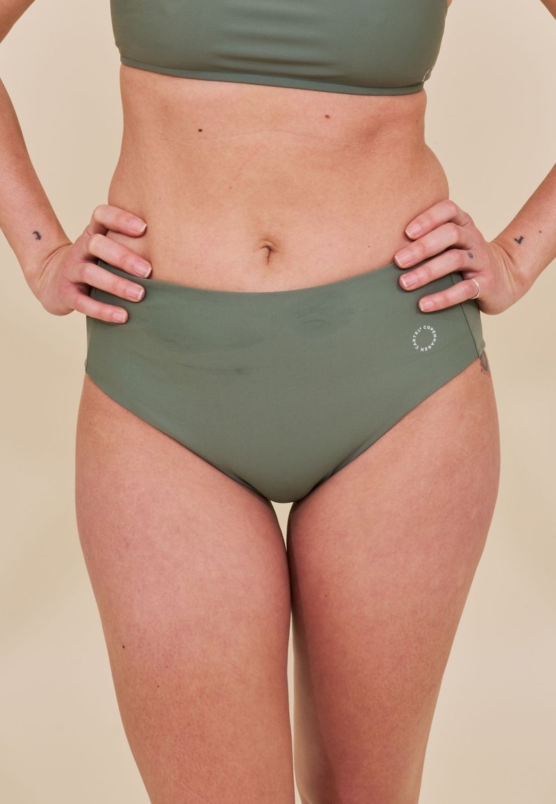 Kobieta w oliwkowych bikini, ręce opierające się na biodrach, beżowe tło, widoczne tatuaże na nadgarstkach.