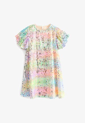 Bambino che indossa un vestito pastello con paillettes e stivali di gomma arcobaleno scintillanti, in piedi sull’erba vicino a striscioni colorati di tessuto e una tenda decorata.