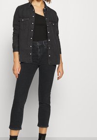 Mujer con chaqueta de mezclilla negra sobre top negro de un solo hombro, jeans skinny de tiro alto negros y botas negras de tobillo contra un fondo liso.