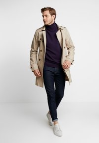 Beige Trenchcoat mit doppelt geknöpftem Vorderteil, lila Rollkragenpullover, dunkle Jeans und weiße Sneaker. Glatte Textur, keine sichtbaren Muster.
