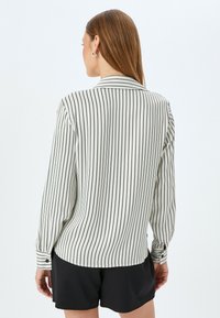 Camisa de manga larga con fondo crema y finas rayas verticales negras, puños con botones y un cuello clásico, combinada con pantalones cortos negros.