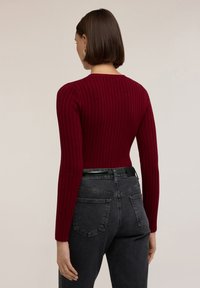 Suéter burdeos acolchado con mangas largas, diseño ajustado y cuello redondo, combinado con jeans oscuros de cintura alta y un cinturón negro.
