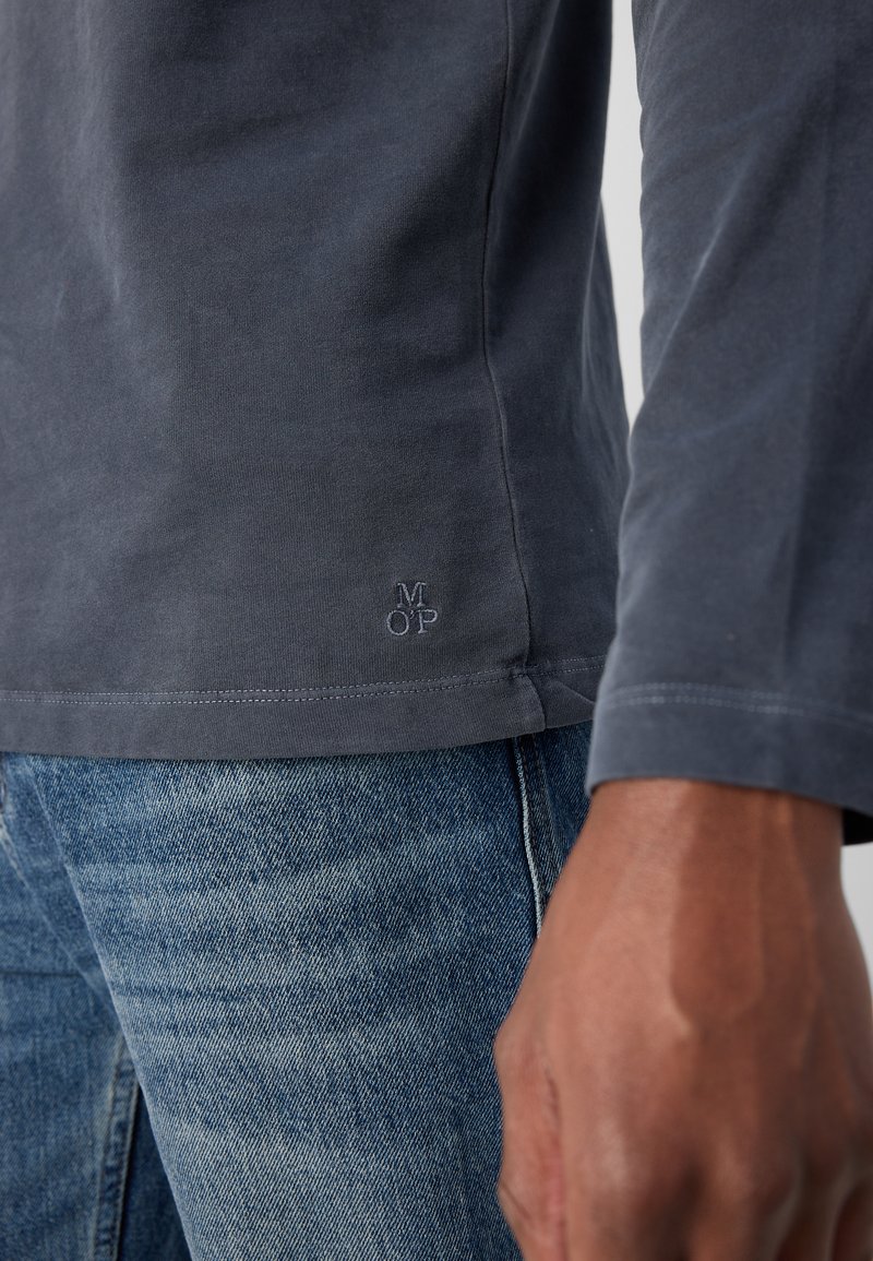 Langärmliges, dunkelgraues Baumwollhemd mit dezentem, gesticktem Logo am Saum. Kombiniert mit hellblauen Jeans.