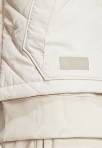 Chaqueta acolchada de color beige claro con dobladillo acanalado y tela texturizada, presenta una etiqueta de logo plateada en el lado izquierdo.