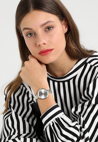 Silverur med rund urtavla, minimalistisk design och guldaccent, bärs på ett handled, tillsammans med en svartvitt randig topp.