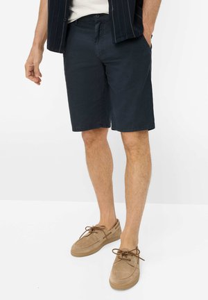 Mand iført navy shorts, beige afslappede snøresko og en mørkstribet skjorte med den ene hånd i lommen.