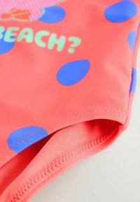 Korall ujumisriiete kangas suurte siniste täppidega, millel on tekst "RAND?" dekoratiivse õmbluse lähedal, lapiku kaeluse ja siledaga.