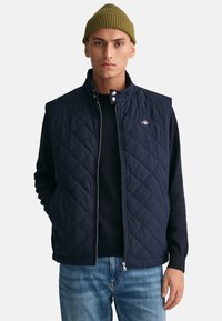 Navy quilted vest with a high collar, zip closure, and snap buttons, worn over a dark sweater. Features a small logo on the left chest.