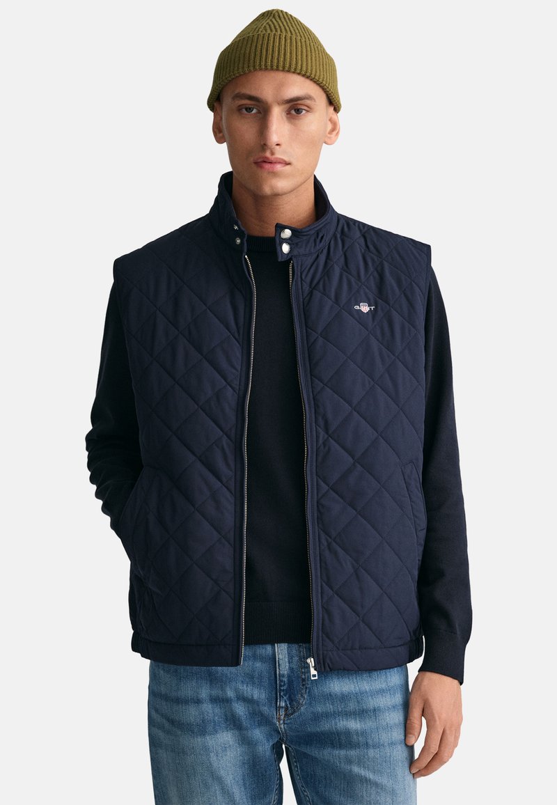 Navy quilted vest with a high collar, zip closure, and snap buttons, worn over a dark sweater. Features a small logo on the left chest.
