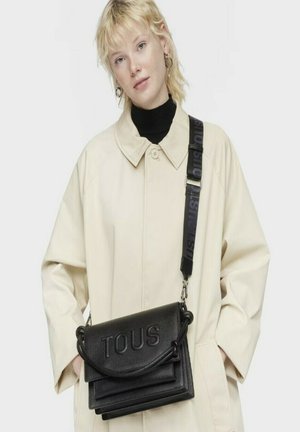 Bolso de piel negro tipo bandolera con el logotipo "TOUS" en relieve, que presenta un acabado texturizado y una correa desmontable con detalles impresos.