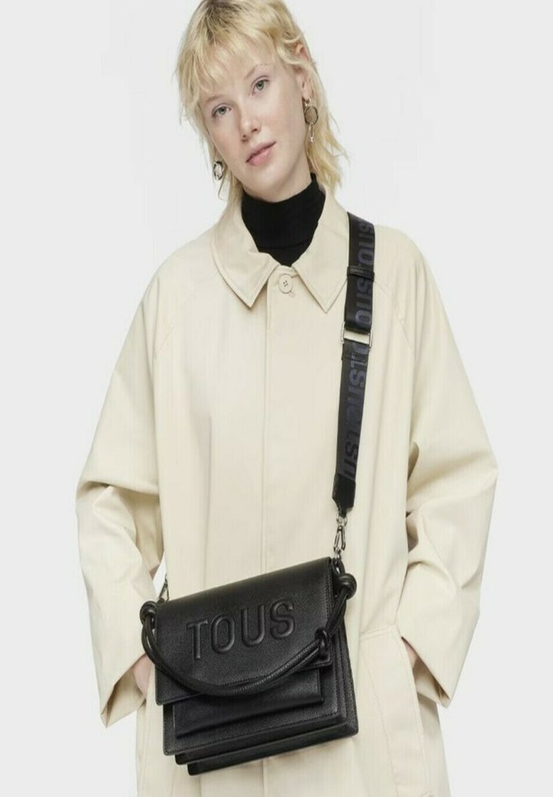 Bolso de piel negro tipo bandolera con el logotipo "TOUS" en relieve, que presenta un acabado texturizado y una correa desmontable con detalles impresos.