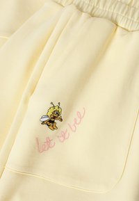 Tissu jaune doux avec une petite abeille de dessin animé brodée et un texte cursif rose indiquant « lot de bee » près du bord d'une poche.