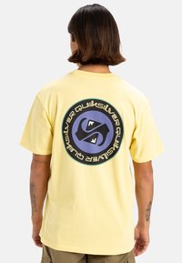 Camiseta de algodón amarilla con un gran gráfico circular en la espalda con un diseño en azul y negro y texto en blanco. Mangas cortas.