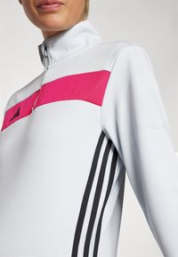 Light grey athletic jacket with a high collar, featuring a pink horizontal stripe across the chest and black three-stripe accents on sleeves.