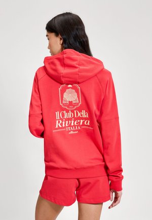 Ženska z dolgimi temnimi lasmi, ki nosi rdečo jopico z kapuco in ujemajoče kratke hlače, na hrbtu jopice je napis "Il Club Della Riviera Italia" s pripadajočo grafiko.