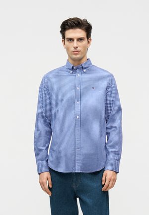 Νέος άνδρας που φορά ένα μπλε και λευκό καρό πουκάμισο με κουμπιά και σκούρο μπλε τζιν, στέκεται μπροστά σε ένα απλό ανοιχτό γκρι φόντο.