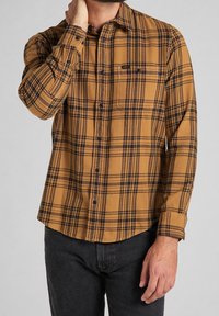 Camicia in flanella a quadri marrone e nera con chiusura a bottone, taschino sul petto, e maniche lunghe, abbinata a jeans scuri.