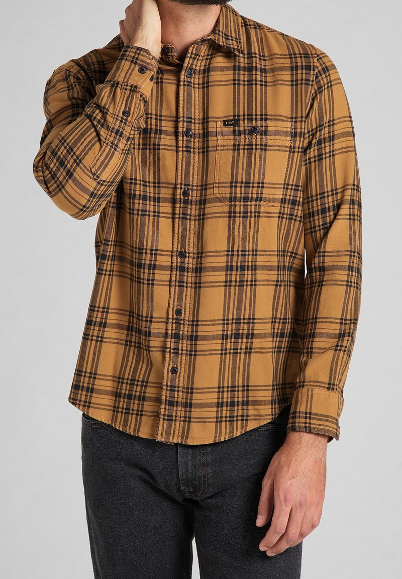 Camicia in flanella a quadri marrone e nera con chiusura a bottone, taschino sul petto, e maniche lunghe, abbinata a jeans scuri.