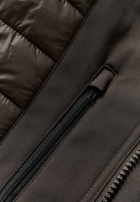 Brun kviltat tyg med en slät, matt yta. Har en svart dragkedja i en dold ficka. Sömmar synliga längs kanterna.