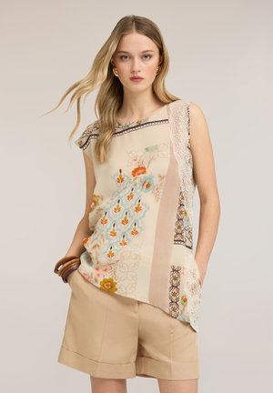 Joven mujer con largo cabello rubio que lleva una blusa sin mangas con estampado floral y pantalones cortos beige con las manos en los bolsillos.