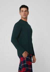 Zielona koszula z długim rękawem, okrągłym dekoltem i dopasowanymi rękawami, w połączeniu z czerwono-granatowymi spodniami do piżamy w kratę z kieszenią.