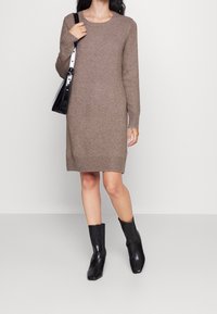 Abito maglione marrone con maniche lunghe, scollatura a girocollo e orlo a coste. Abbinato a stivali neri alla caviglia e una borsa a tracolla nera.