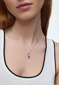 Srebrny naszyjnik z sercowym wisiorkiem, który ma centralny różowy kamień otoczony przez przejrzyste akcenty, wyeksponowany na białej bluzce.