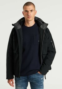 Chaqueta blanda negra con capucha, cierre frontal con cremallera, dos bolsillos en el pecho y un interior texturizado. Se lleva sobre un suéter azul marino y unos vaqueros azules.