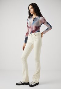 Bluză lungă, cu mâneci, din material transparent, cu un model abstract colorat; asortată cu blugi albi, cu talie înaltă și pantofi cu accente metalice masive.