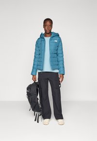 Teal pufferjakke, lyseblå topp, svarte cargo-bukser, kremfargede sko. Svart ryggsekk med stropper, teksturerte materialer og tettsittende design.