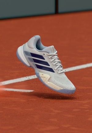 Chaussure de tennis blanche avec une tige en mesh, des accents bleus et trois bandes contrastantes. Conception légère adaptée aux surfaces de terrain.