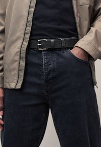 Jeans di denim nero con taglio dritto, indossati con una camicia scura e una giacca grigio chiaro con zip. Una cintura di pelle nera presenta una fibbia argentata.