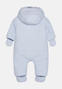 Combinaison de ski rembourrée bleu clair avec capuche, poignets élastiques et chaussons intégrés. Design matelassé texturé, avec des accents navy aux manches.