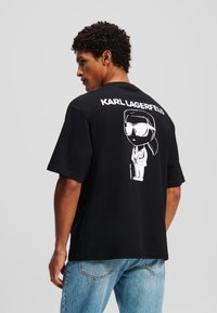Fekete, rövid ujjú póló, fehér grafikával, amelyen napszemüveget viselő figura látható, a tervezés fölött pedig a "KARL LAGERFELD" felirat.