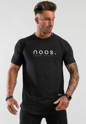 Man med stylat hår och tatueringar som bär en svart "noos luxury basics" t-shirt och en svart smartklocka, tittar ner och åt vänster.