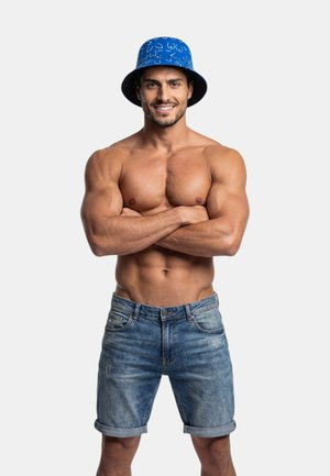 Muskularny mężczyzna ze skrzyżowanymi ramionami, noszący niebieski kapelusz typu bucket z wzorem oraz podwinięte krótkie spodenki z denimu, uśmiechający się na tle gładkiego tła.