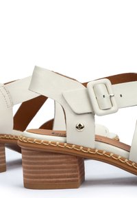 White leather sandals with a chunky wooden heel, featuring cross straps, a square buckle closure, and decorative stitching along the sole.