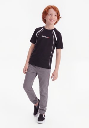 Rød håret dreng smiler og kigger sidelæns, iført en sort Quiksilver t-shirt, grå bukser og sorte slip-on sko.