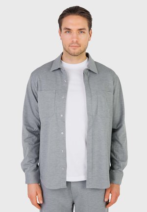 Ung mand iført en lysegrå skjorte med knapper over en hvid t-shirt, stående mod en ensartet lys baggrund, vendt mod fronten.