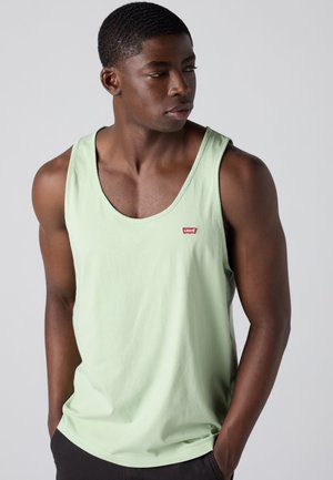 Młody mężczyzna o ciemnej karnacji ubrany w jasnozielony top tank Levi's, patrzący na bok na tle jednolitego szarego tła.