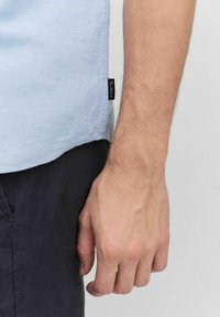 Lichtblauw kortemouwen shirt van linnen, met een afgeronde hem en een zwart stoffen label aan de zijkant, gecombineerd met donkere broeken.