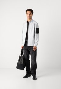 Jachetă butonată gri deschis cu accente negre, tricou negru, pantaloni din denim închis și pantofi negri. Poartă o geantă neagră.