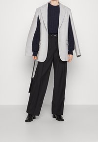 Blazer ample gris clair porté sur un pull en maille torsadée bleu marine, associé à un pantalon noir taille haute et des bottines noires. Accessoires : un petit sac noir.