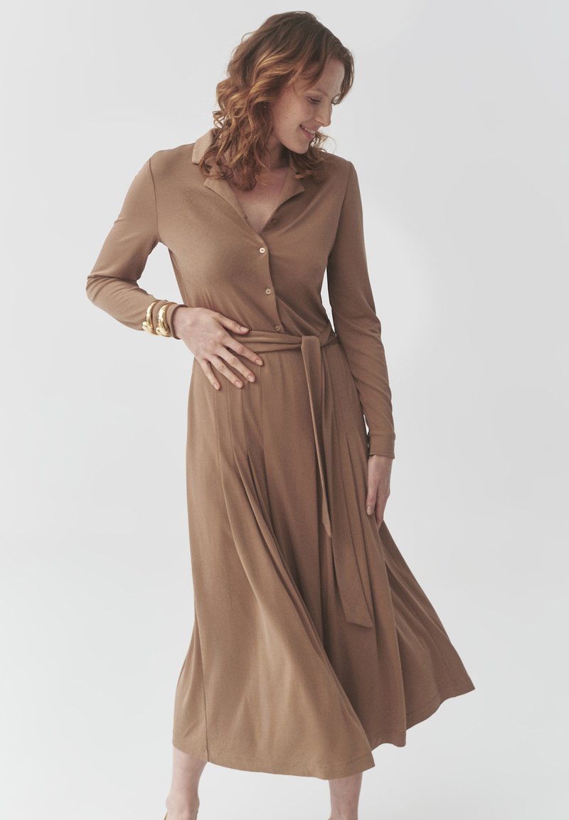 Braunes, langärmliges Kleid mit Kragen, Knopfleiste vorne und einem Bindegürtel in der Taille. Fließender Rock mit Falten, aus weichem Stoff.