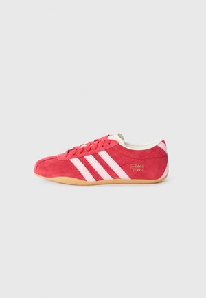 Red suede athletic shoe with pink stripes, lace-up closure, and cream-colored accents. Features a gum sole and embroidered branding.