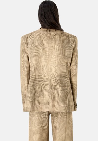 Personne aux longs cheveux foncés portant un blazer beige à carreaux surdimensionné et un pantalon large assorti, vue de dos.