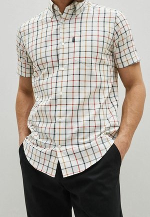 Man wearing short-sleeve button-up shirt with white base and thin black, brown, and red plaid lines, paired with black pants.